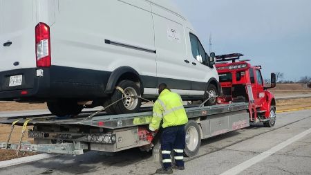 White panel van on flatbed tow truck with worker in yellow jacket